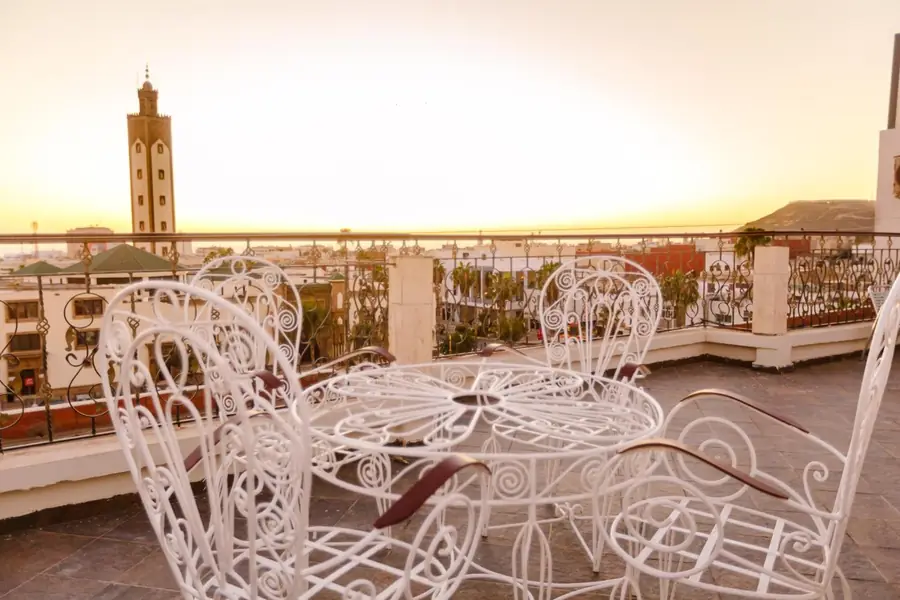The rooftop terrace of Hôtel Lynx at sunset: white wrought-iron chairs, the minaret of the Mohammed V Mosque on the left and the Kasbah hill on the right.