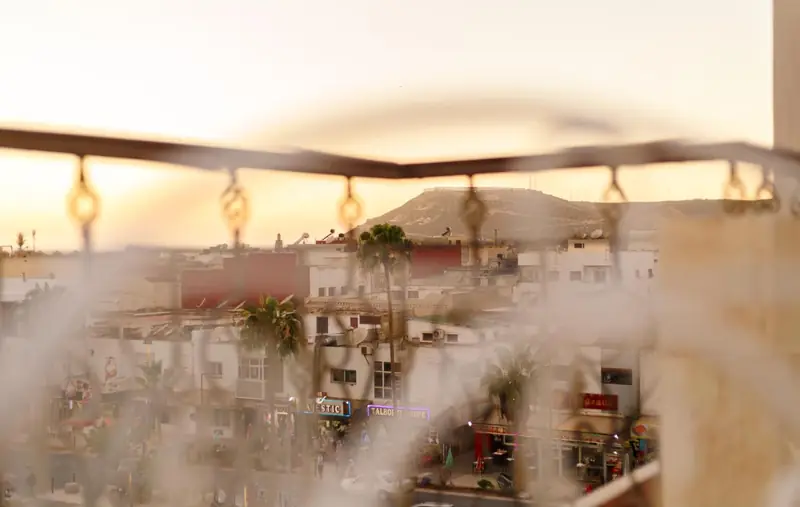 Through the blurred iron rail of a Hôtel Lynx balcony: Talborjt rooftops, palm trees, lit shops and the Kasbah hill behind.